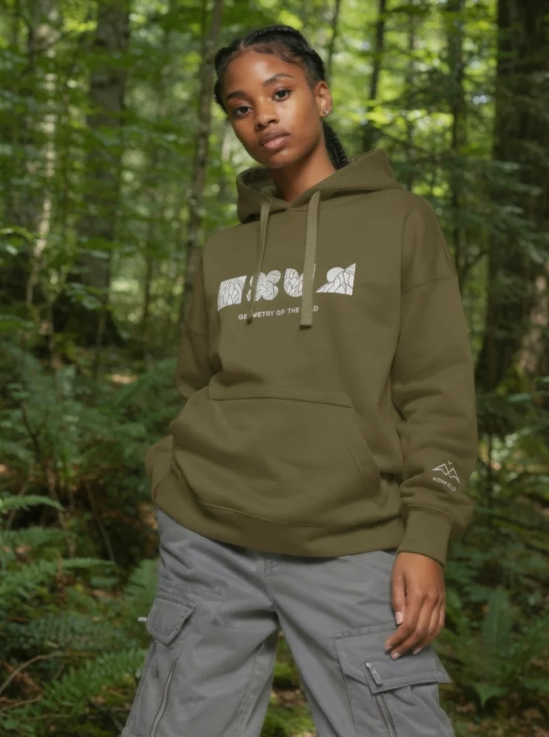 Geometry of the Wild Military Green Hoodie by NOWFIELD porté dans un paysage naturel, illustrant l'esprit outdoor et minimaliste de la collection.
